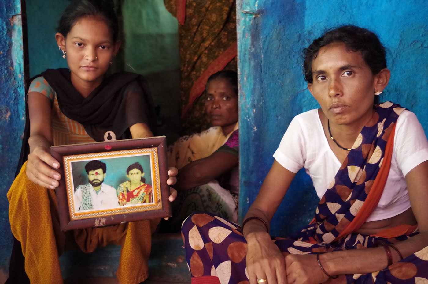 Three women sitting in the doorway of a house. One is holding a framed photograph of her deceased father with her mother.