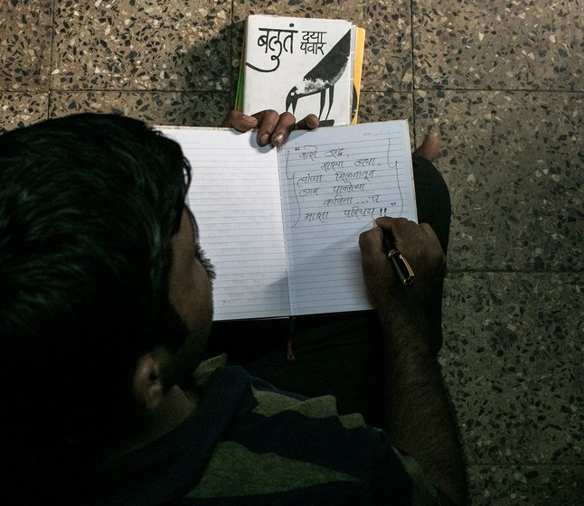 A man sitting on the floor and writing in a book. A copy of the book 'Baluta' is lying next to him