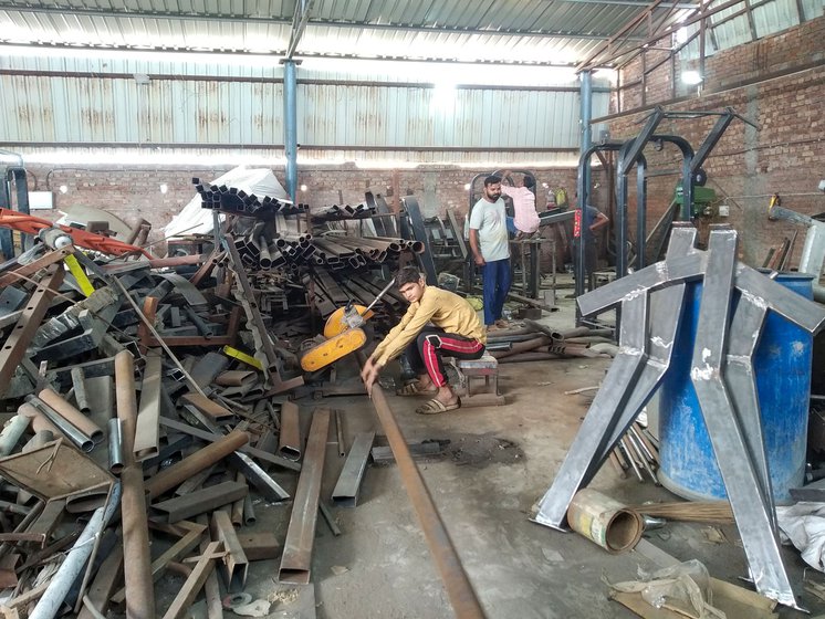 Asif pushes the iron pipe along the empty floor on his left to place it on the cutting machine; he cuts (right) the 15 feet long iron pipe that will go into making the 8 station multi-gym