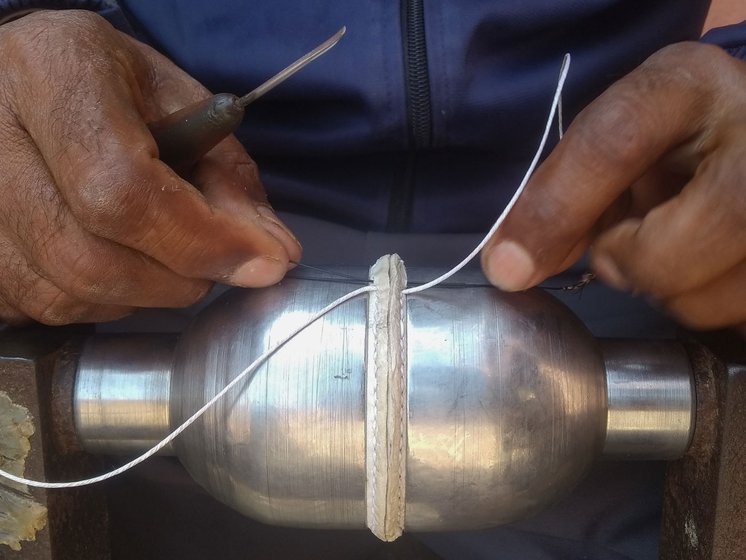 Bharat Bhushan using an aar to make insertions through the leather that protrudes between the two hemispheres, held together by an iron clamp. He places a rounded cork between the two cups and attaches pig bristles by their roots to the ends of a metre-long cotton thread for the second stage of stitching. He then inserts the two pig bristles through the same holes from opposite directions to stitch the cups into a ball